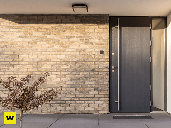 Moderne Haustür aus Aluminium neben heller Klinkerfassade mit kleinem Baum im Eingangsbereich.